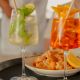 Two cocktail glasses on a black tray with a shrimp and lemon plate between them: a pale mojito-like drink with lime and mint on the left, and an orange Aperol-style drink with orange slices on the right.