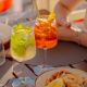 Two colorful cocktails on a wooden patio table—one lime-mint drink and one orange cocktail with a slice of orange—beside a plate of fried seafood and lemon wedges.