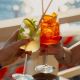 Two cocktails are raised for a toast: a clear lime-mint drink and a bright orange citrus drink, clinking over a sunlit outdoor table by the water.