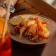 Plate of fried seafood with a lemon wedge on a wooden table, with a tattooed arm and a red drink visible in the foreground.