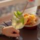 Hand holding a lime-mint cocktail on an outdoor table, with a blurred plate of food and lemon wedges in the background.