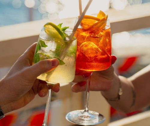 lariva_beachbar_sunset Two people clink cocktail glasses outdoors, with lime and mint garnishes on a sunlit table.